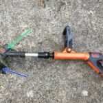 A person inspecting a broken cordless strimmer in a sunny green garden.